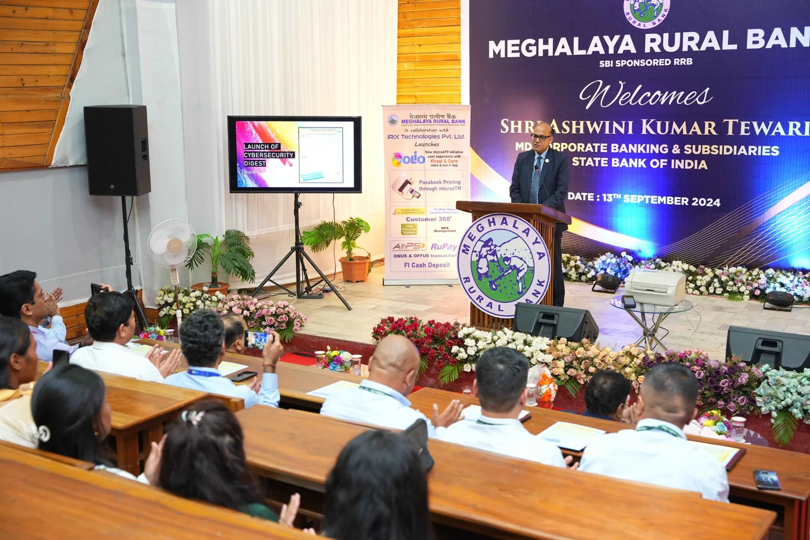 Shri Ashwini Kumar Tewari, MD (Corporate Banking & Subsidiaries), State Bank of India, speaking at the Meghalaya Rural Bank event on 13 September 2024, beside a banner announcing OLiO Passbook Printing through microATM in collaboration with IRIX Technologies